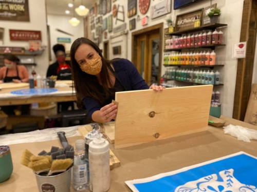 A woman in a mask works on a wooden board at a crafting workshop, surrounded by art supplies and colorful paint.