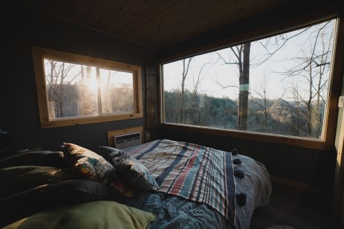 Cozy bedroom with a bed, colorful pillows, and large windows showcasing a serene forest view at sunrise.