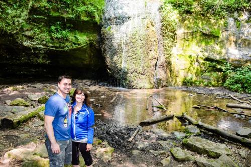 A couple stands smiling by a serene waterfall surrounded by lush greenery and a calm pond.