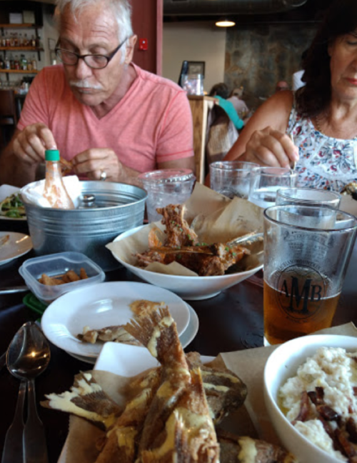 A dining scene with two people enjoying a meal, featuring plates of fried fish, salad, and drinks on the table.