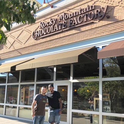 Two men stand outside the Rocky Mountain Chocolate Factory, smiling in front of the shop's entrance.