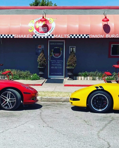 Two sports cars, one red and one yellow, parked in front of a diner with a retro sign and outdoor seating.