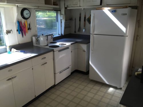 Bright kitchen with white cabinets, a refrigerator, stove, and a sink, featuring sunlight streaming through the windows.