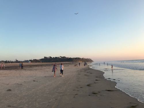People walking along a sandy beach at sunset, with gentle waves and a clear sky in the background.