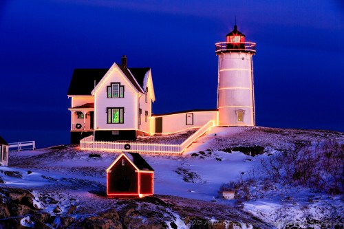 A lighthouse and house illuminated with festive lights, surrounded by snow under a twilight sky.