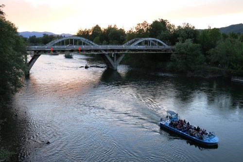 A boat with passengers glides on a river beneath a bridge at sunset, surrounded by trees and mountains.