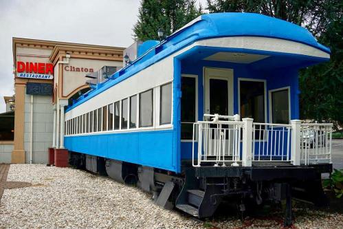 A blue diner built from a vintage train car, with large windows and a welcoming entrance, surrounded by gravel and greenery.