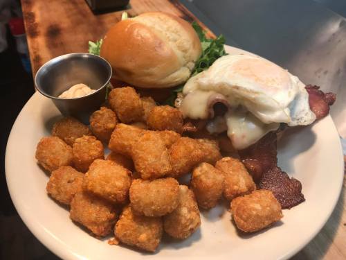 A plate with a sandwich topped with an egg and bacon, served with tater tots and a small cup of mayonnaise.