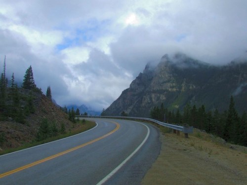 A winding road curves through a mountainous landscape under a cloudy sky, surrounded by trees and rocky terrain.