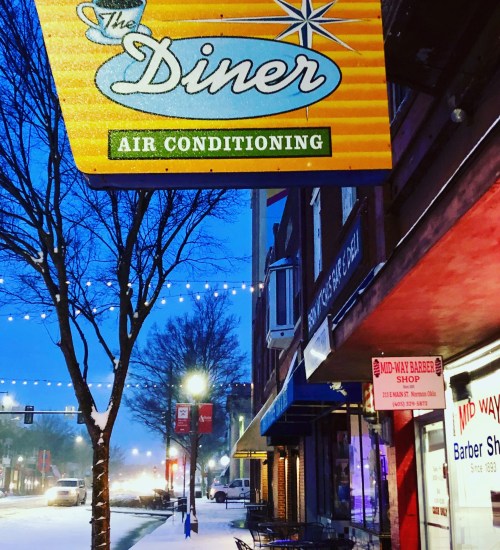 A diner sign hangs above a snowy street, with trees and buildings illuminated by soft lights in the evening.