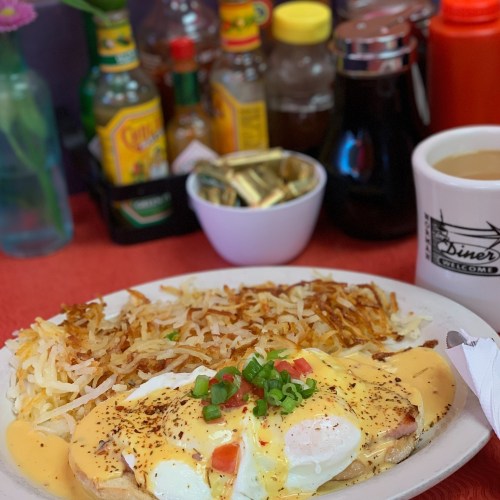 A plate of eggs Benedict with hash browns, garnished with green onions, alongside condiments and a coffee cup.