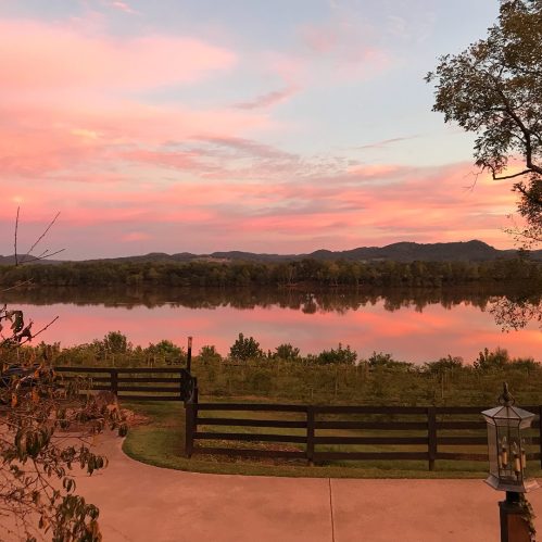 A serene landscape at sunset, featuring a calm river reflecting pink and orange hues, surrounded by lush greenery and hills.