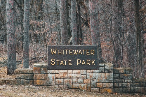 Sign for Whitewater State Park surrounded by bare trees and a rocky stone base.