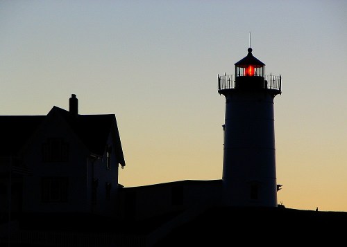 Silhouette of a lighthouse at sunset, with a glowing red light and a nearby house in shadow.