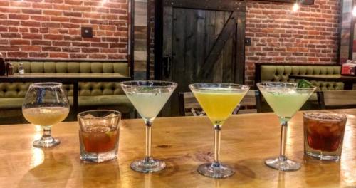 A wooden bar with five colorful cocktails in glasses, set against a rustic brick wall and green upholstered seating.