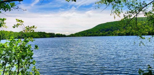 A serene lake surrounded by lush green trees and hills under a partly cloudy sky.