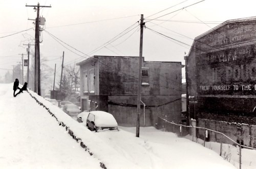A snowy street scene with a person sitting on a snowbank, buildings, and a car covered in snow. Power lines overhead.