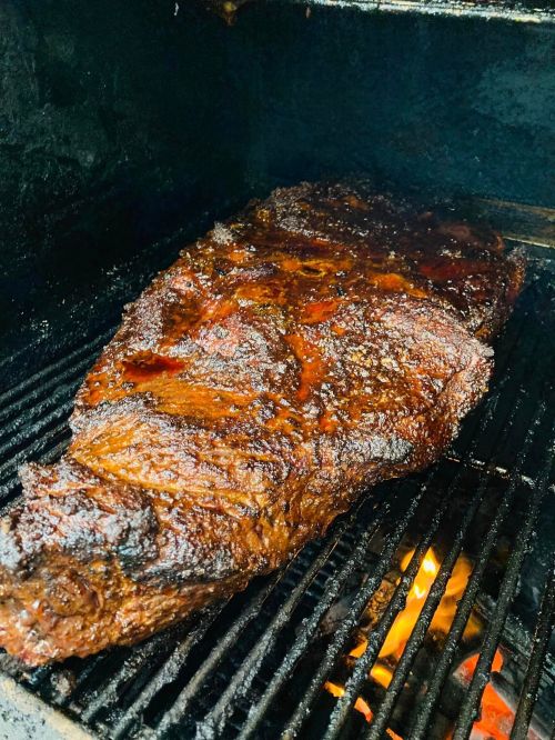 A large, smoked brisket resting on a grill, with a dark, caramelized crust and glowing embers below.