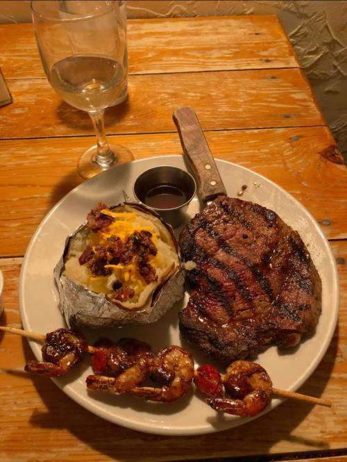 A plate with a grilled steak, shrimp skewer, baked potato topped with cheese and bacon, and a glass of white wine.