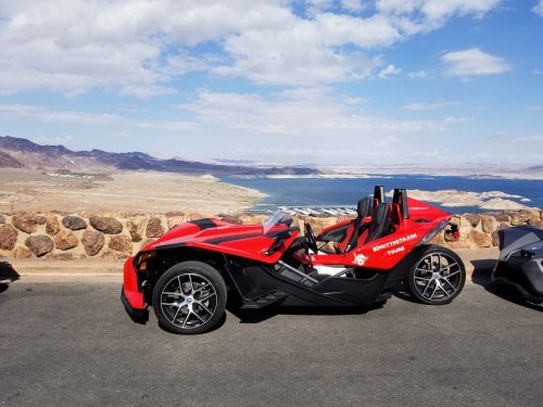 A red three-wheeled vehicle parked by a scenic overlook with mountains and a lake in the background.