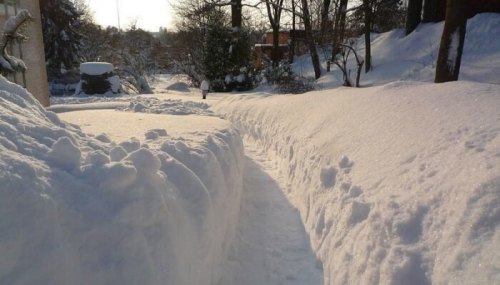 A snow-covered pathway surrounded by high snowdrifts, with trees and a distant building in the background.