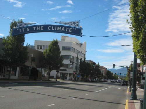 A street view of Grant's Pass with a sign reading "IT'S THE CLIMATE" and trees lining the road under a blue sky.
