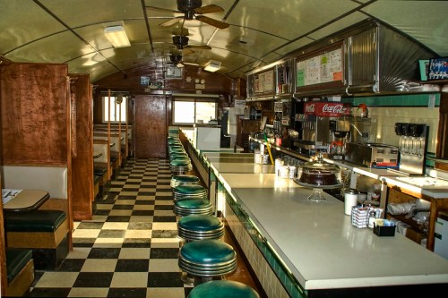 Interior of a vintage diner with checkered floors, a long counter, and booth seating, featuring retro decor and a nostalgic vibe.