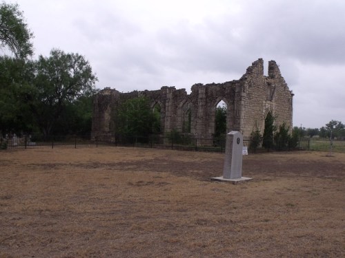 Ruins of a stone building with arched windows, surrounded by grass and trees, under a cloudy sky.