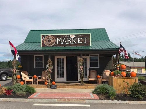 A quaint market with a green roof, pumpkins, and flags, featuring a welcoming porch with wooden chairs.