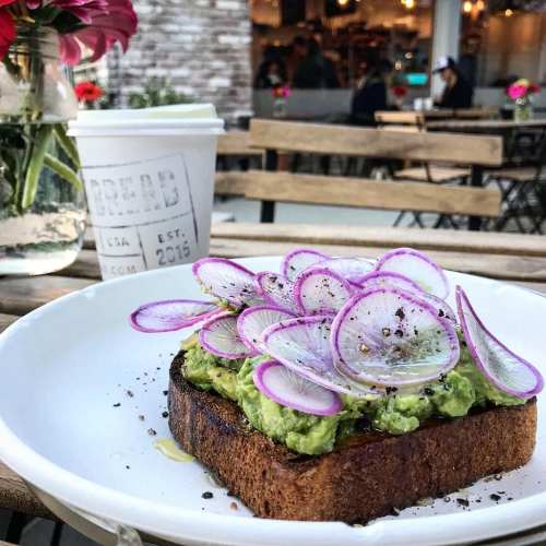 A plate of avocado toast topped with purple radish slices, served with a coffee cup and flowers in the background.