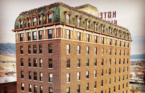 Historic brick building with a green-tiled roof and a vintage "Hotel" sign on top, set against a city backdrop.