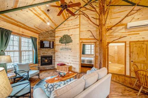 Cozy cabin interior with wooden walls, a stone fireplace, and a tree feature, featuring a living area and bedroom doorway.