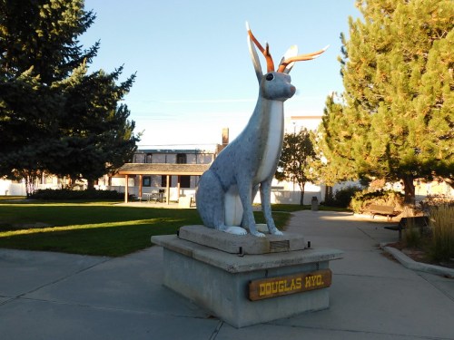 A large statue of a jackrabbit sits on a pedestal in a park, surrounded by trees and a building in the background.