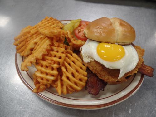 A plate with a fried chicken sandwich topped with a sunny-side-up egg, served with waffle fries, tomato, and pickles.