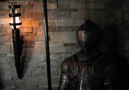 A suit of medieval armor stands against a stone wall, illuminated by a vintage wall sconce.