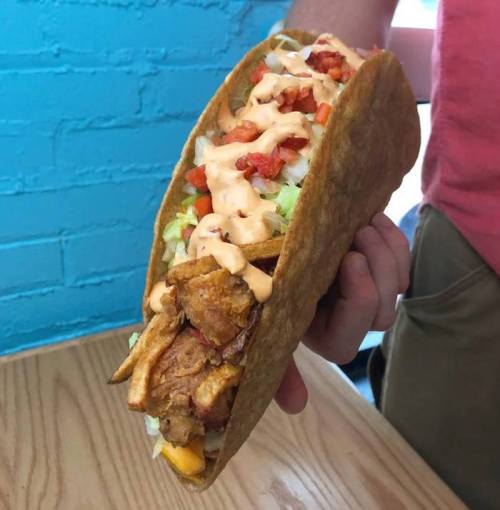 A person holding a large taco filled with fried chicken, lettuce, tomatoes, and drizzled with sauce.