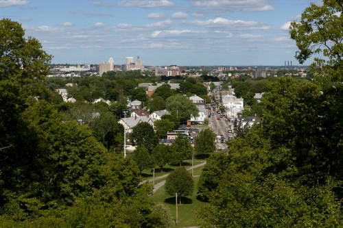 A panoramic view of a city skyline surrounded by greenery and residential areas under a partly cloudy sky.
