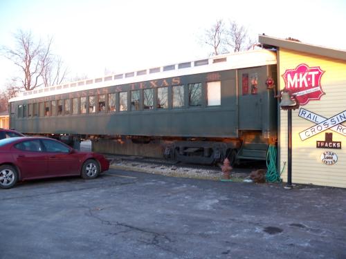 A vintage green train car parked beside a yellow building with a railroad crossing sign.