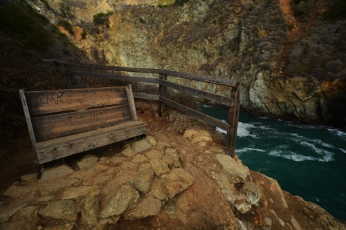 A weathered wooden bench by a rocky cliff overlooking a turquoise river, surrounded by rugged terrain.
