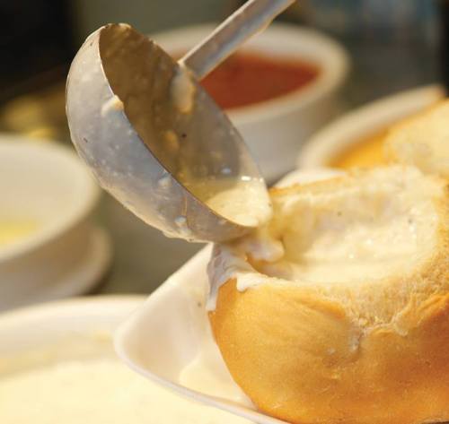 A ladle pouring creamy sauce into a bread bowl, with bowls of sauce in the background.