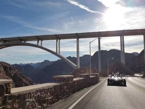 A scenic road curves beneath a large bridge, with mountains in the background and a vehicle driving ahead.