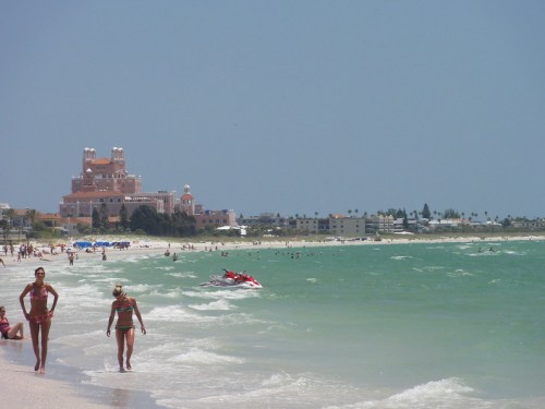 Two women walk along a sandy beach with a resort in the background and people enjoying the water.