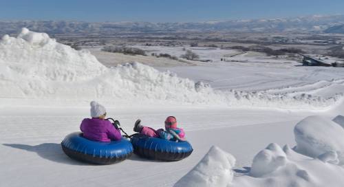 Two children in snow tubes slide down a snowy hill, surrounded by a winter landscape and mountains in the background.