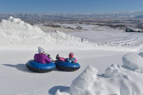 Two people in colorful winter gear sledding down a snowy hill on blue inner tubes, with a scenic snowy landscape in the background.