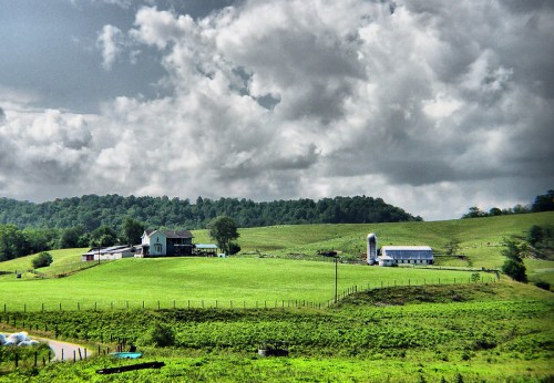 A scenic farm landscape with a house, barn, and silo surrounded by lush green fields under a cloudy sky.