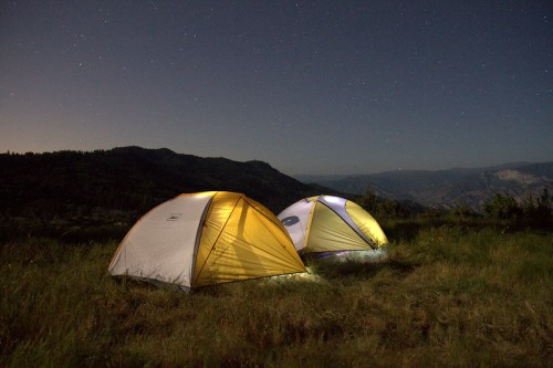Two tents illuminated at night, set against a starry sky and mountains in the background.