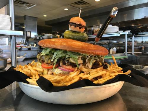 A giant burger topped with a small burger and pickle, surrounded by a mound of fries, on a restaurant counter.