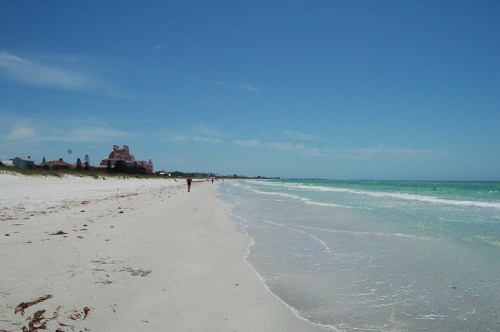 A serene beach scene with soft white sand, gentle waves, and a clear blue sky, featuring a distant figure walking along the shore.