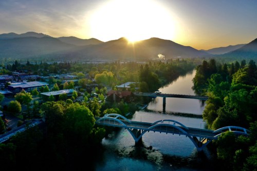 Aerial view of a river with a bridge, surrounded by trees and mountains, as the sun rises in the background.