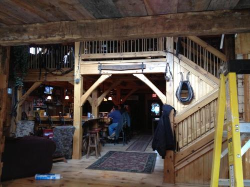 Interior of a rustic bar with wooden beams, a staircase, and patrons seated at the bar. Cozy atmosphere with warm lighting.
